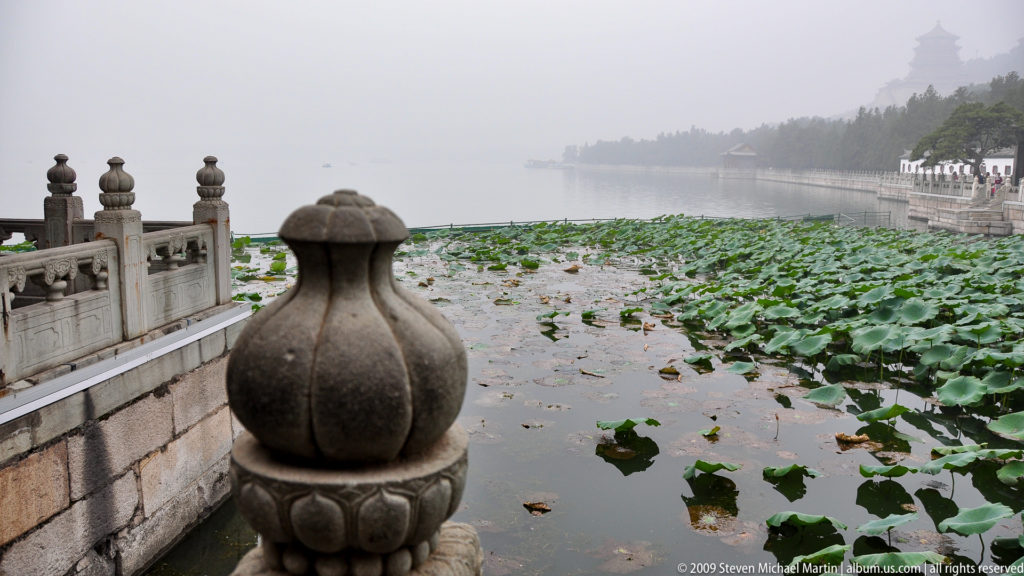 Vase-shaped stone balustrade finial beside dense lotus leaves on hazy Kunming Lake, with a causeway receding along the shore.