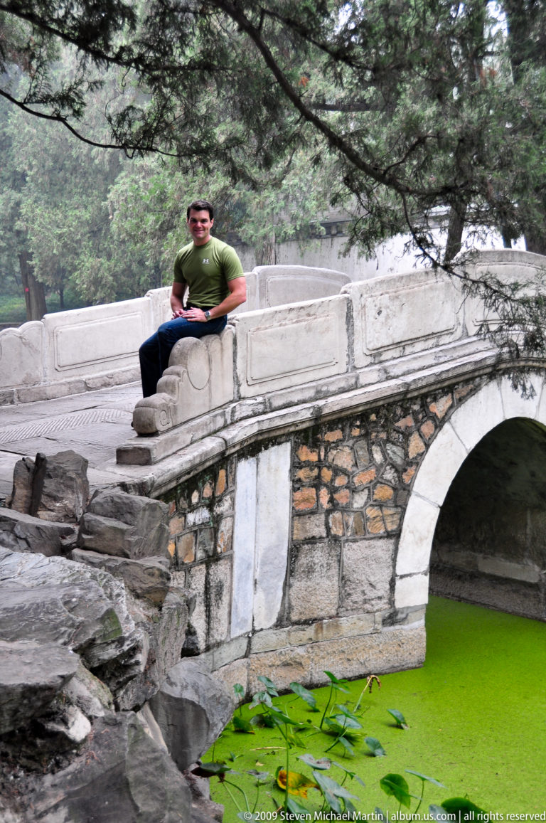 Man seated on the parapet of a small stone arch bridge above bright green pond water and lotus leaves.