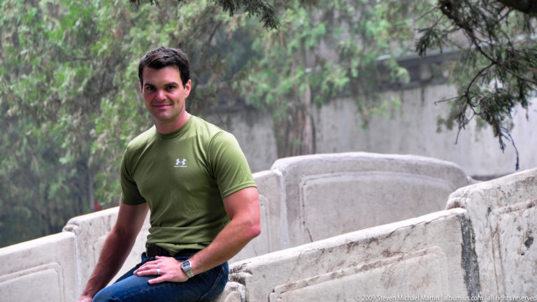 Man in a green shirt seated on a pale carved stone balustrade beneath hanging willow branches at the Summer Palace.
