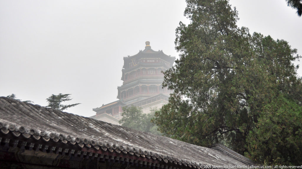 Upper stories of the Tower of Buddhist Incense emerging through haze above gray tiled roofs and trees at the Summer Palace.