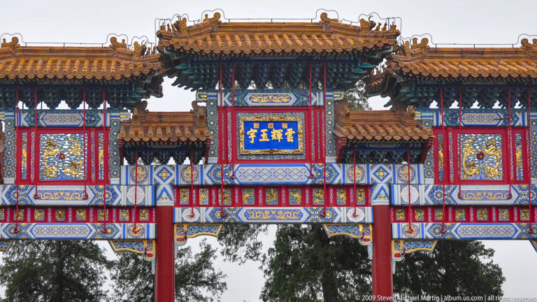 Close view of a richly painted paifang with yellow tiled roofs, red columns, dragon panels, and a blue plaque with gold Chinese characters.