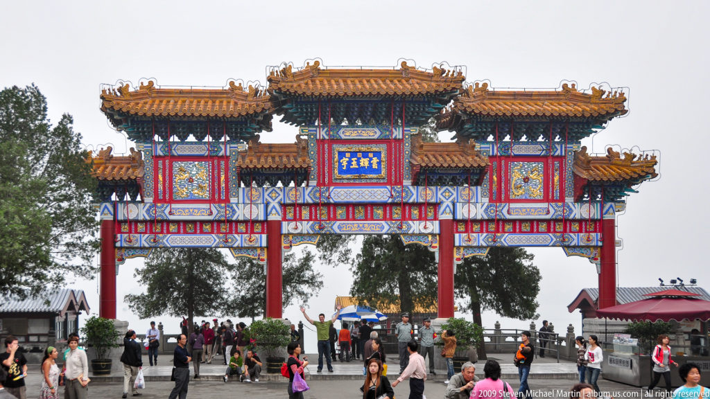 Large colorful Chinese paifang with red columns, yellow tiled roofs, blue-green painted ornament, and visitors passing underneath.