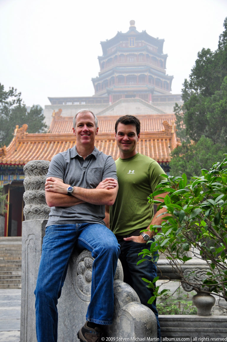 Two men posing at a carved stone railing with orange palace roofs and the hazy Tower of Buddhist Incense rising behind them.