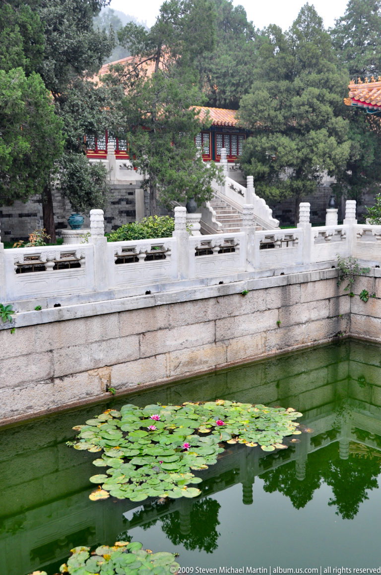Lotus-filled green pond bordered by pale carved stone railings and stairs, with trees and red-roofed buildings beyond.