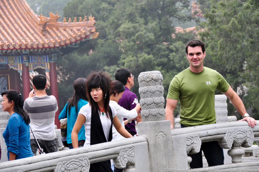 Man in a green shirt standing beside a carved stone balustrade, with visitors, a pavilion roof, and wooded hillside behind him.