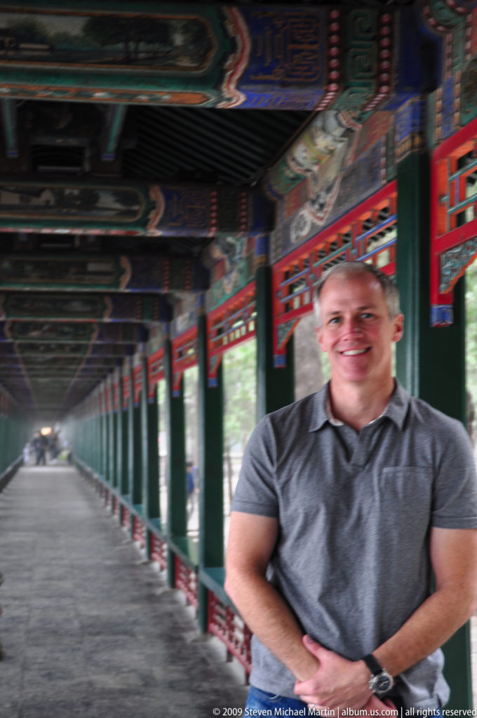 Long painted covered corridor receding behind a seated visitor, with repeating green columns, red railings, and decorated ceiling beams.