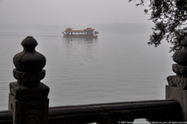Distant covered boat on misty Kunming Lake, framed in the foreground by dark stone balustrade finials and a lakeside railing.
