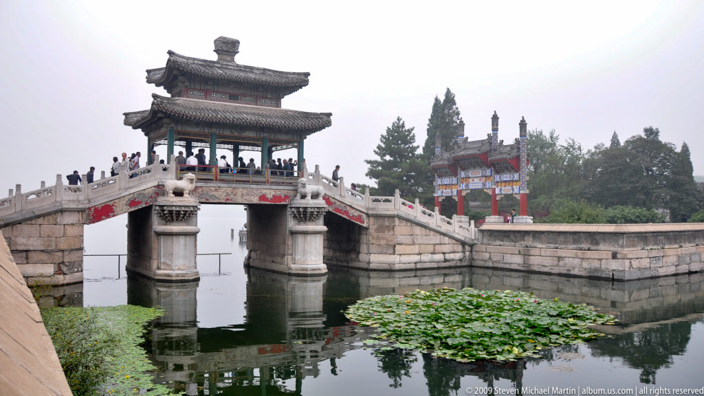 Roofed stone bridge reflected in water beside dense lotus leaves, with a colorful gate and mature trees beyond.