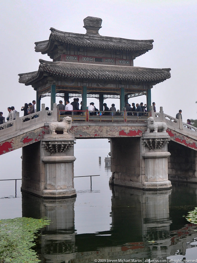 Side view of a stone bridge on masonry piers supporting an open pavilion with layered gray tiled roofs above lotus-covered water.