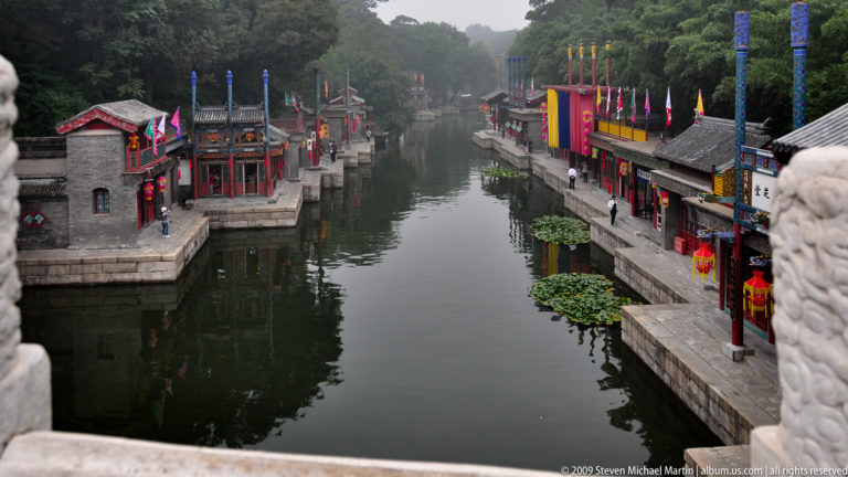 Narrow canal viewed from a bridge, lined by stone embankments, traditional buildings, trees, and colorful banners at the Summer Palace.