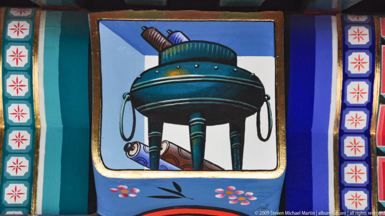 Painted blue architectural panel with a stylized tripod bronze vessel, floral sprigs, and red-and-white geometric borders.