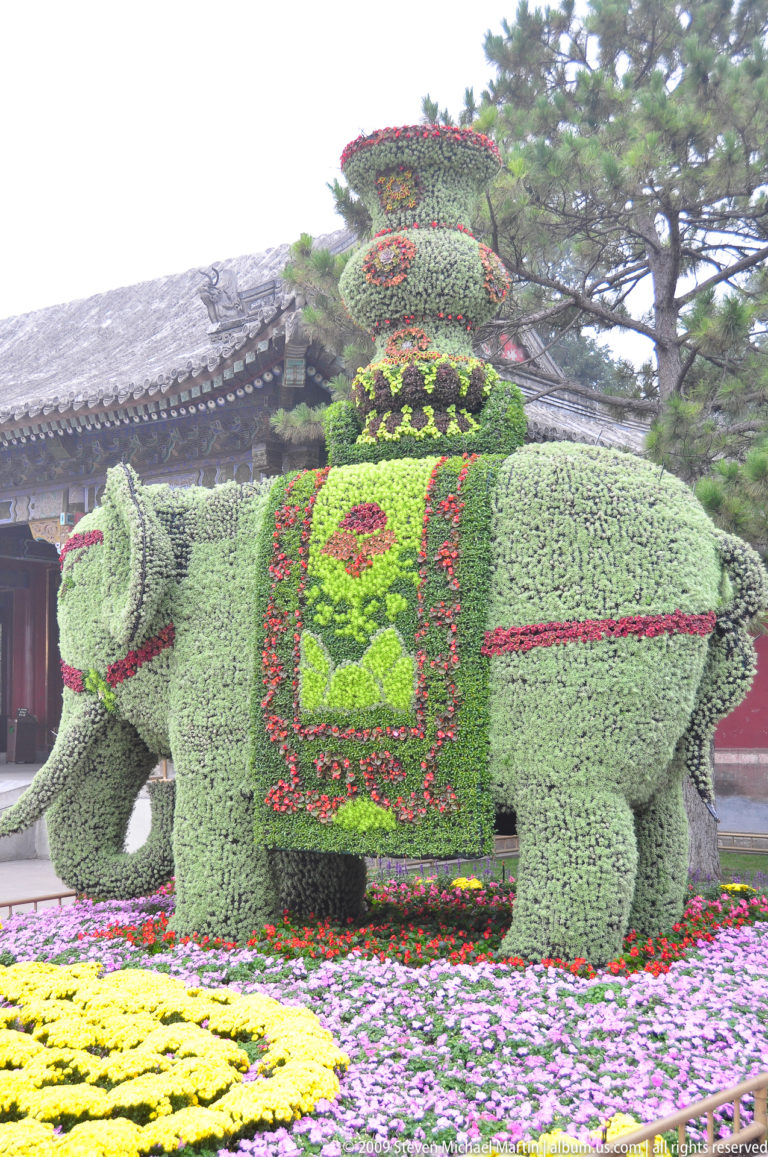 Side view of a large green elephant-shaped floral display with red patterns, tiered back ornament, and colorful flower beds.