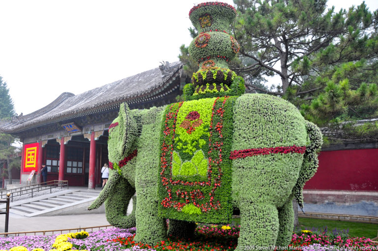 Large elephant-shaped floral sculpture with green foliage, colorful patterns, and a tiered ornament, standing before traditional buildings and trees.