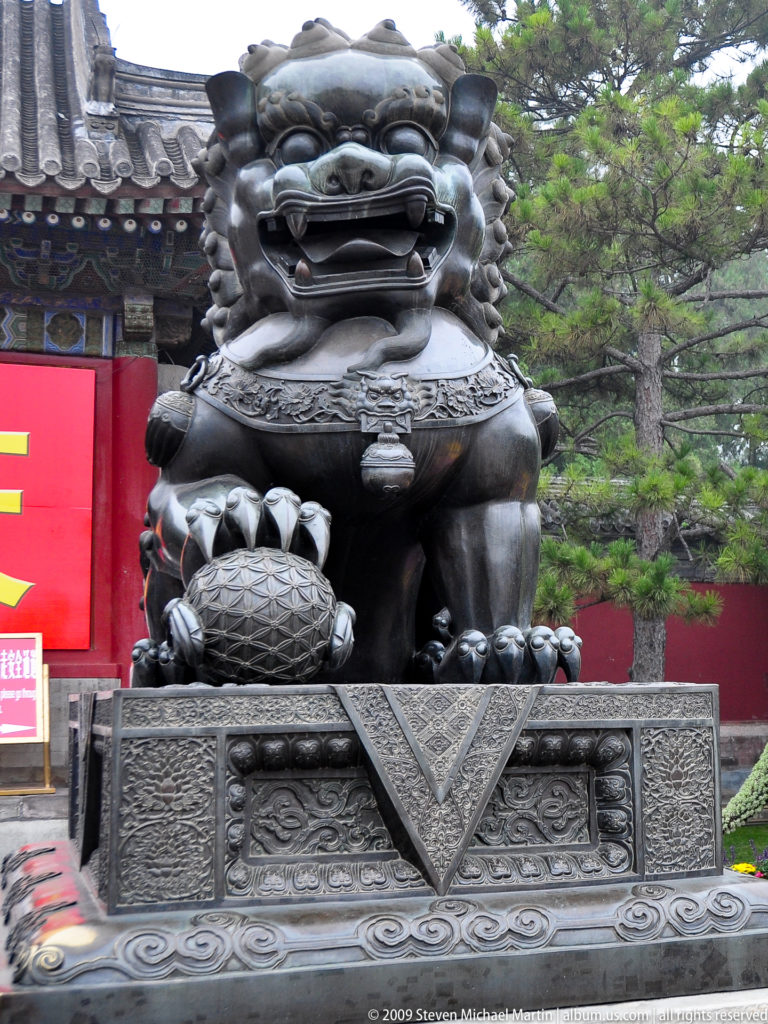 Frontal dark stone guardian lion on an ornate pedestal, with one paw resting on a patterned ball and trees behind.
