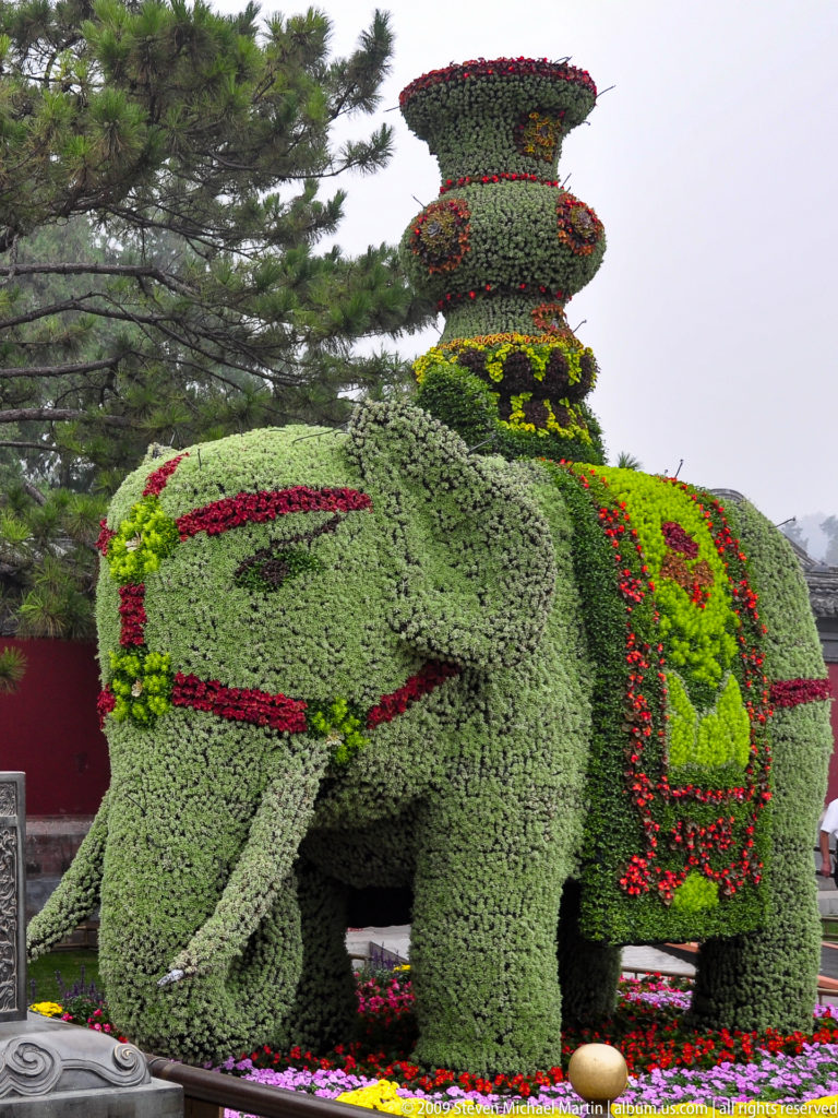 Large elephant-shaped topiary covered in green foliage and red floral bands, with a tall tiered planted ornament on its back.