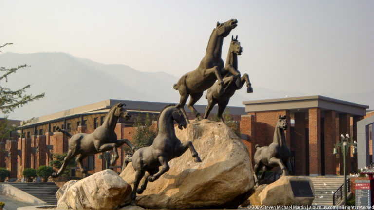 Modern monumental sculpture of dark-metal galloping horses outside the Mausoleum of the First Qin Emperor / Terracotta Army museum complex near Xi'an, with museum buildings and distant mountains in the background.