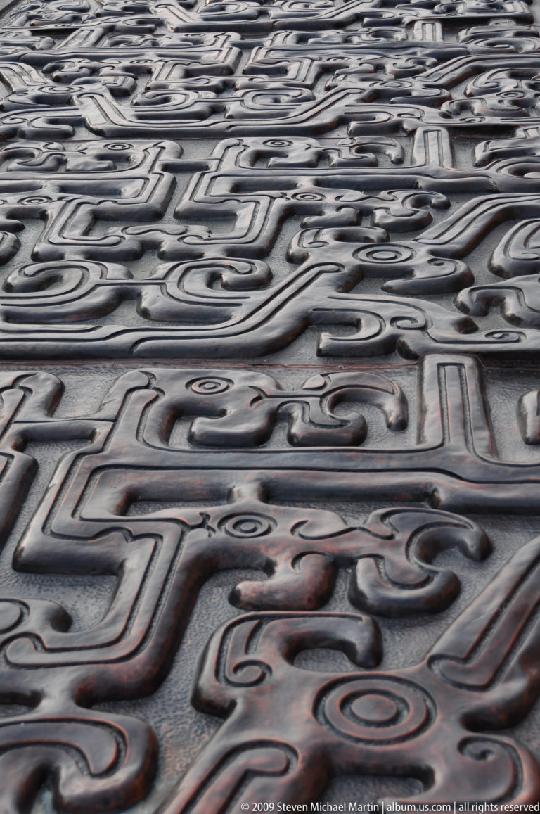 Close-up of interlocking scrolls and mask-like forms in archaizing relief on cast bronze paving.