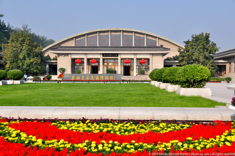 Symmetrical main exhibition hall at the Terracotta Army museum complex near Xi’an, with a broad arched roof and colonnaded entrance behind red and yellow flowerbeds.