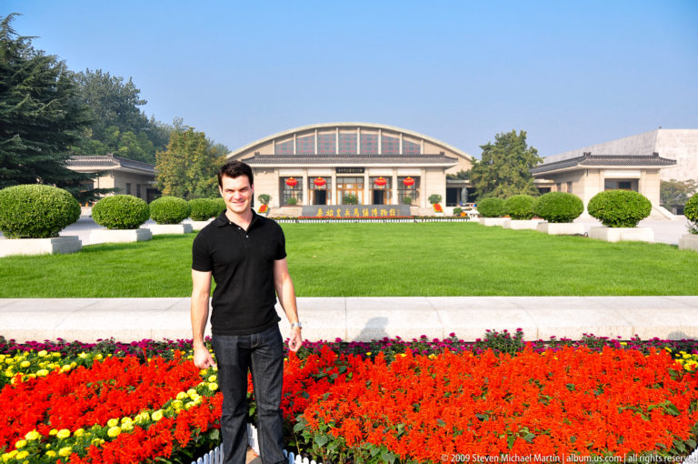 Full-length portrait of a male visitor in dark clothing standing on the landscaped approach in front of the arched main exhibition hall at the Terracotta Army museum complex near Xi'an.