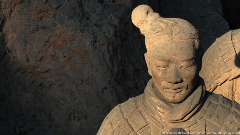 Close portrait of an armored terracotta warrior from the Mausoleum of the First Qin Emperor, showing his modeled facial features, topknot, and rectangular chest armor plates.