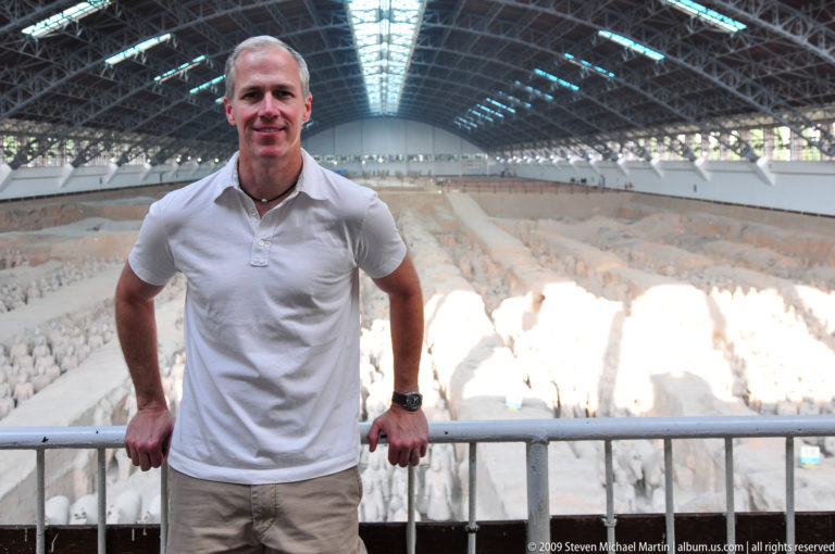 Male visitor in a white shirt at the railing above Pit 1 with rows of terracotta warriors and the steel-roofed excavation hall behind him.