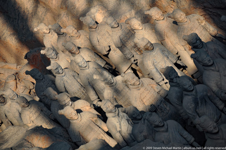 Oblique overhead view of tightly packed terracotta infantry figures standing in ordered rows under strong light and shadow.