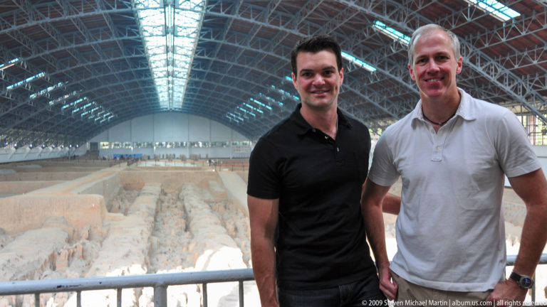 Two male visitors stand at a railing overlooking Pit 1 of the Terracotta Army, with rows of terracotta warriors and the large hall roof visible behind them.