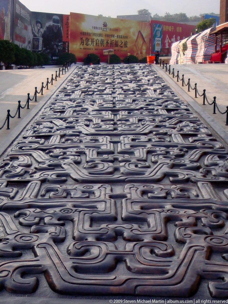Close view of dark cast bronze paving panels decorated with symmetrical scroll and mask-like motifs.