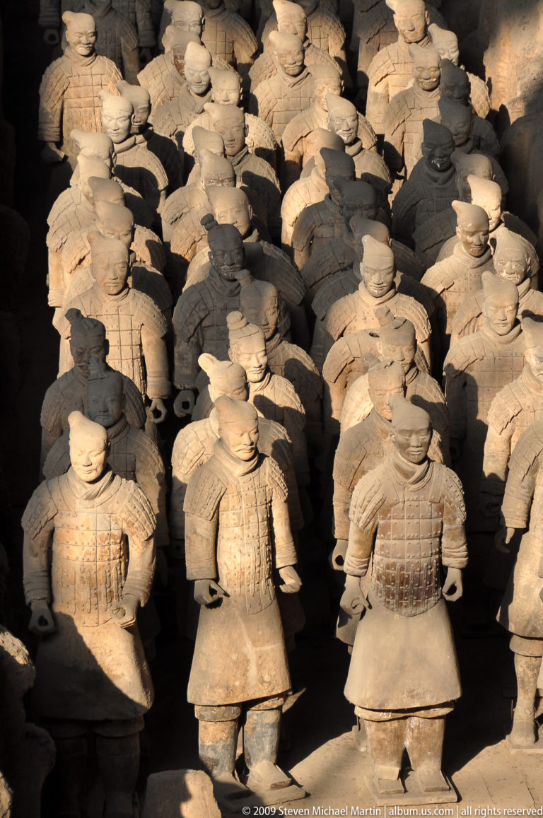 Front row of standing terracotta infantry warriors under raking light, their upper bodies illuminated while lower areas fall into shadow.