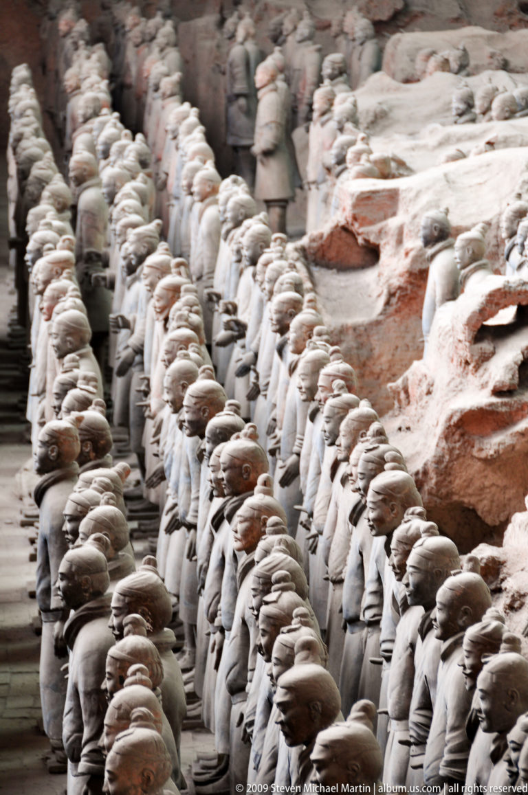 Side view of long rows of terracotta infantry soldiers in tightly aligned ranks along an earthen wall.