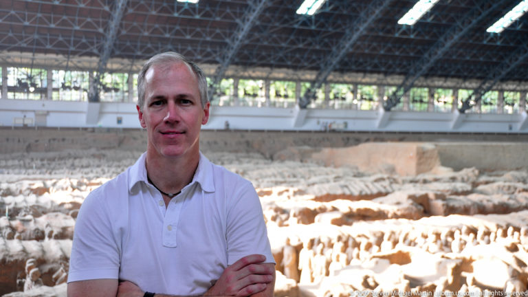 Man in a white polo shirt stands with folded arms in front of Pit 1 at the Terracotta Army museum, with rows of terracotta warriors and the steel-roofed hall visible behind him.