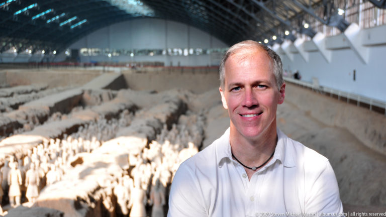 Smiling male visitor in a white polo shirt standing beside the excavation, with terracotta warriors and earthen trenches blurred in the background.