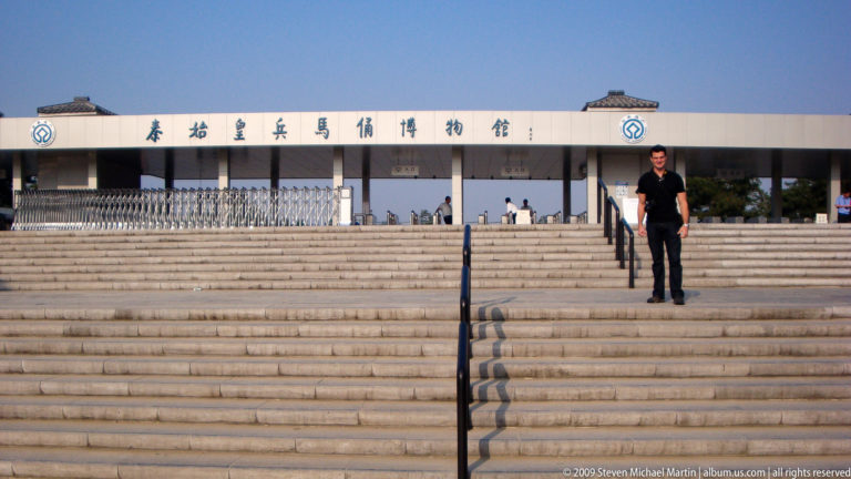 Broad stairway leading up to the modern entrance of the Museum of the Terracotta Warriors and Horses, with a visitor on the steps beneath a clear blue sky.