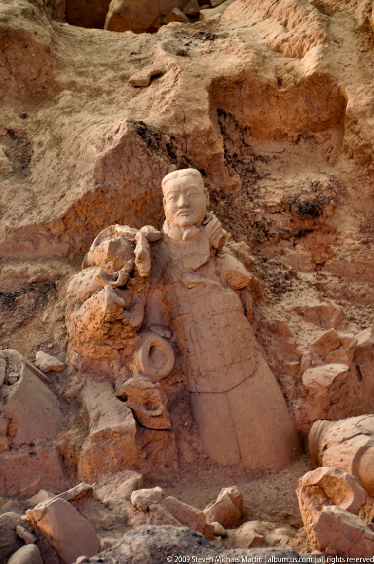 Damaged Qin dynasty terracotta warrior with one arm missing, standing against an earthen background.