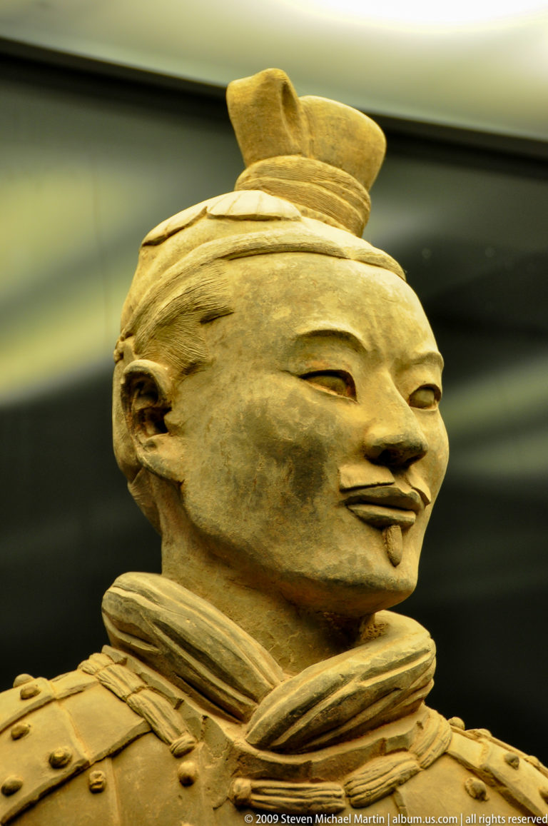 Close-up of a museum-displayed terracotta warrior head from the Qin dynasty, showing preserved facial features including arched brows, a moustache, and modeled lips against a dark background.
