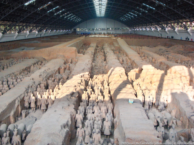 Wide overview of Pit 1 at the Terracotta Army site, showing rows of excavated terracotta warriors in earthen corridors under a large steel-framed museum hall.