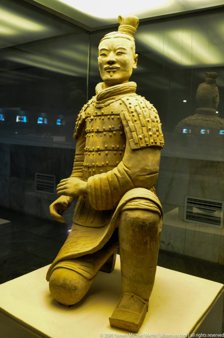 Lower torso and bent knee of a terracotta kneeling armored archer behind glass in a museum display.
