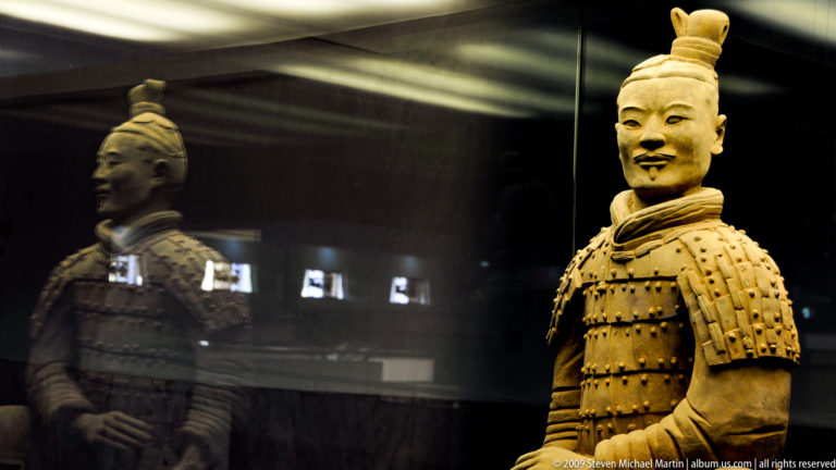 Terracotta armored warrior with moustache and tied hair standing behind glass in museum display.