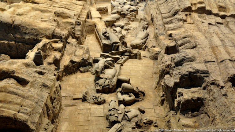 Collapsed terracotta warriors and horse fragments lying across a brick-paved excavation corridor with earthen walls at the Terracotta Army site near Xi’an.