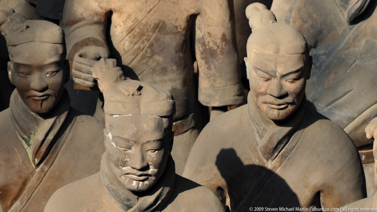 Close view of damaged and intact terracotta warrior heads, with a broken foreground head overlapping a larger sunlit moustached figure behind.