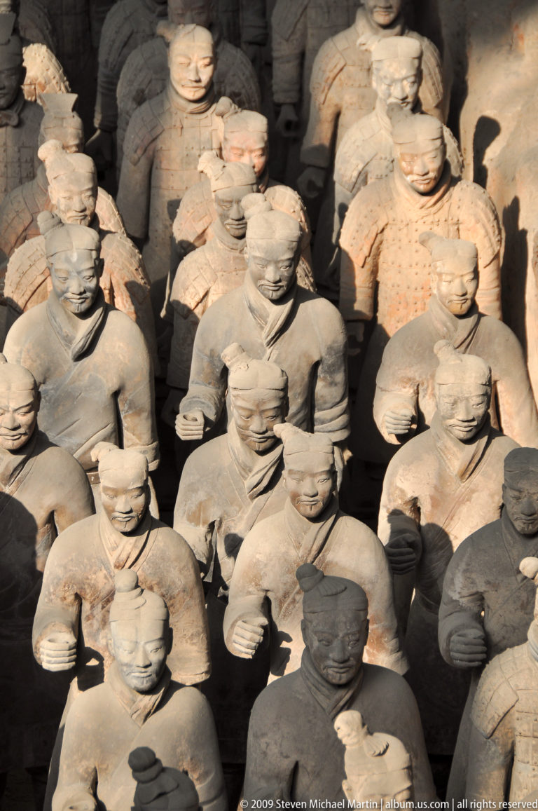 Rows of robed, unarmored terracotta infantry figures with distinct moustaches and hairstyles standing closely packed together.