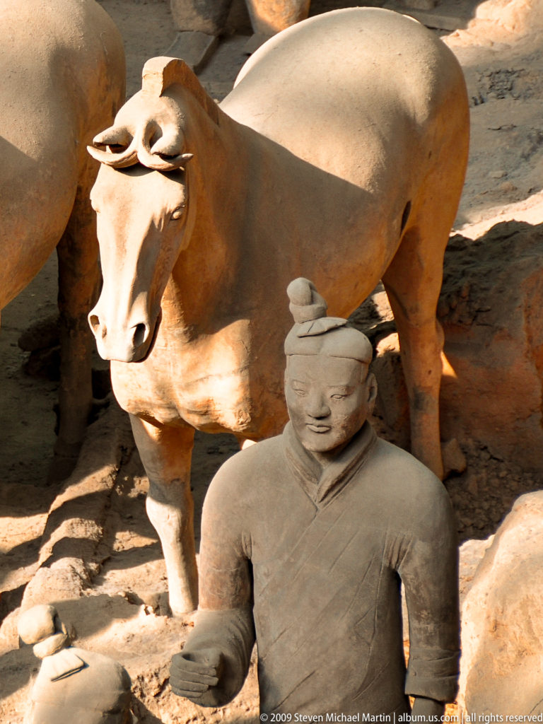 Close view of a terracotta horse’s muzzle and forelegs behind a standing robed warrior figure from the Qin dynasty Terracotta Army.