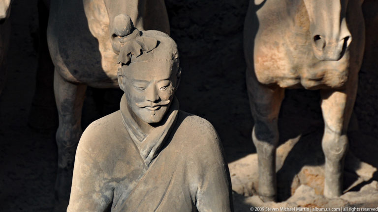Close view of a robed terracotta warrior with a damaged head and moustache, with part of a terracotta horse visible in the background.