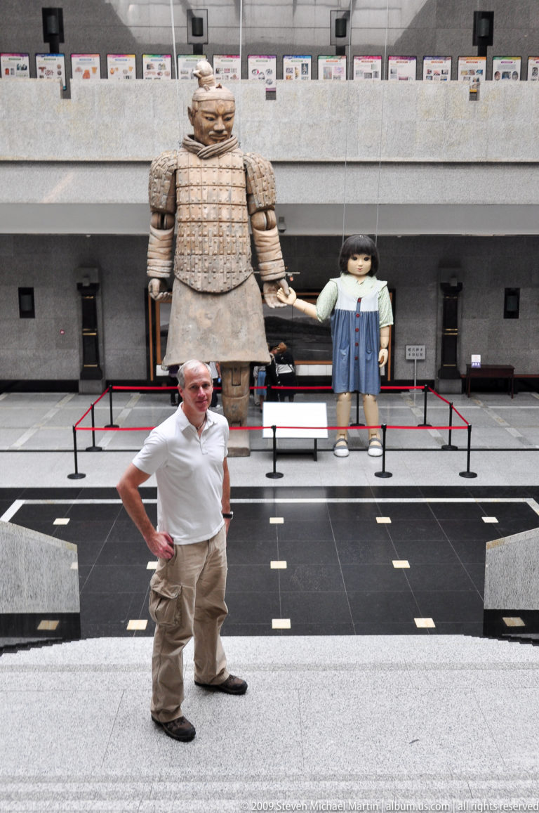 Male visitor posing in a museum with an oversized terracotta-warrior figure and child mannequins behind red stanchions.