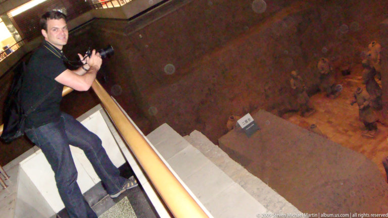 Visitor leaning over a railing to look down into a deep earthen excavation shaft at the Terracotta Army museum complex near Xi'an.