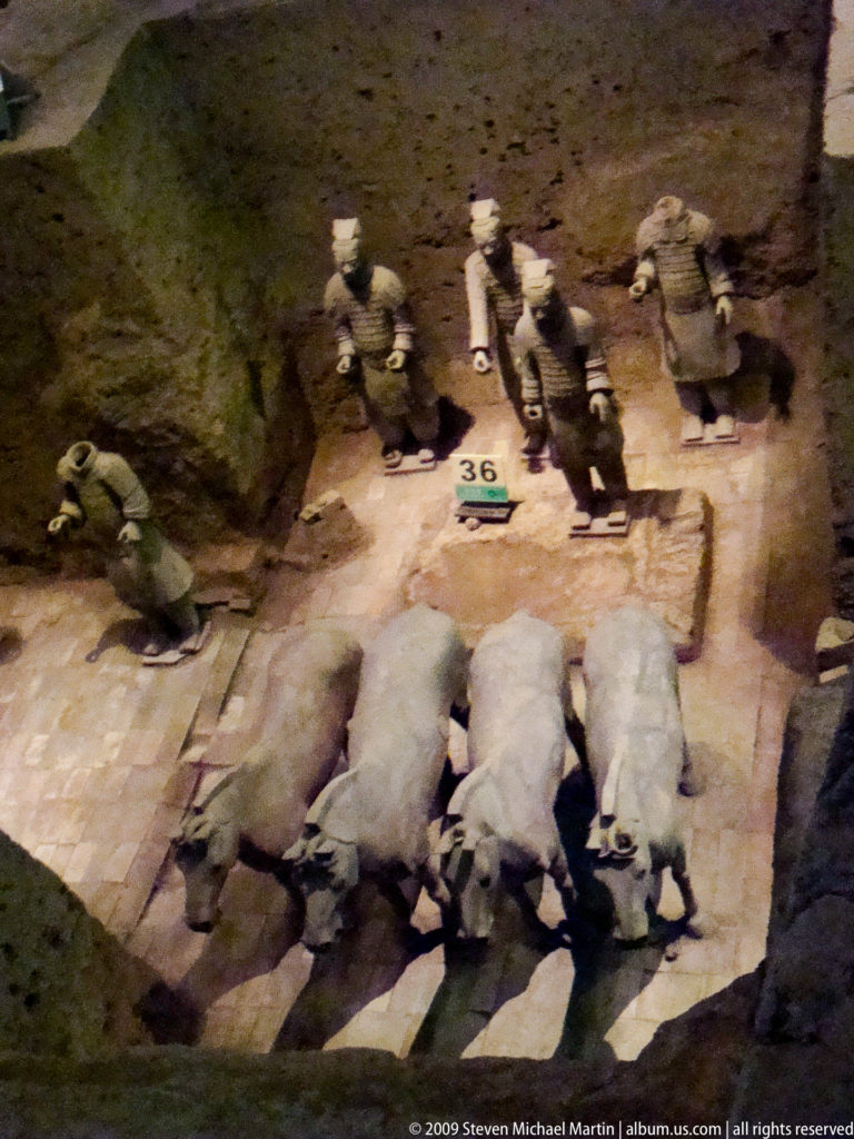 Three excavated terracotta horses aligned side by side with standing warrior figures behind them at the Terracotta Army site near Xi'an.