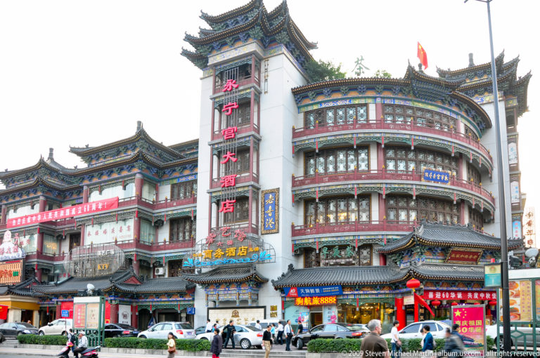 Curved corner hotel in Xi’an with ornate traditional-style Chinese roofs and a vertical red 大酒店 sign facing a busy street with cars and pedestrians.