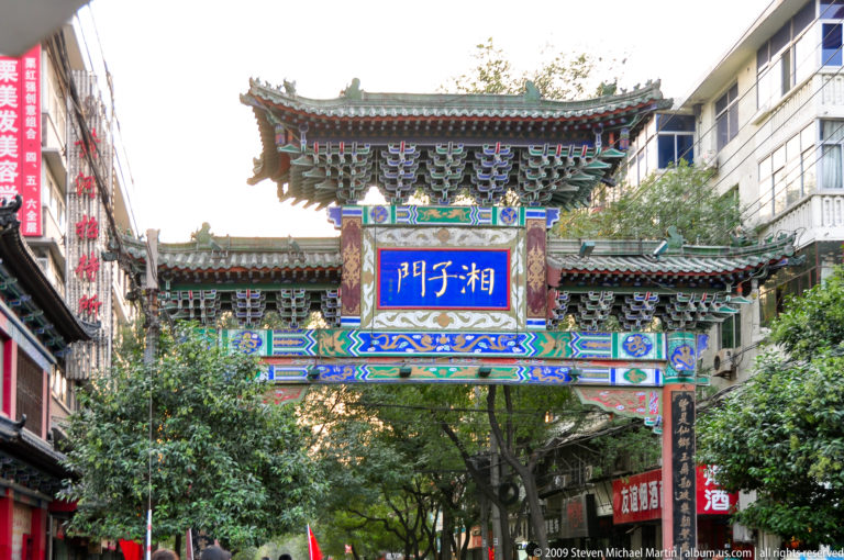 Street view of the Xiangzi Gate (湘子门) paifang spanning a narrow, tree-lined commercial lane with shops in Xi’an.