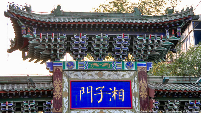 Close view of the ornate Xiangzi Gate (湘子门) paifang in Xi’an, showing colorful dougong brackets and a blue inscribed plaque under a tiled roof.