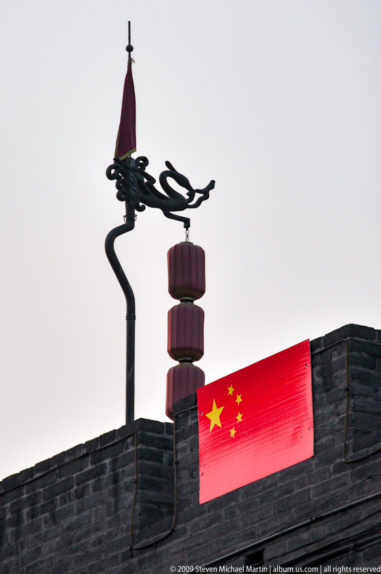 Chinese flag with one large and four smaller yellow stars flying beside three stacked red lanterns on a dragon-shaped street bracket against a pale sky in Xi’an.